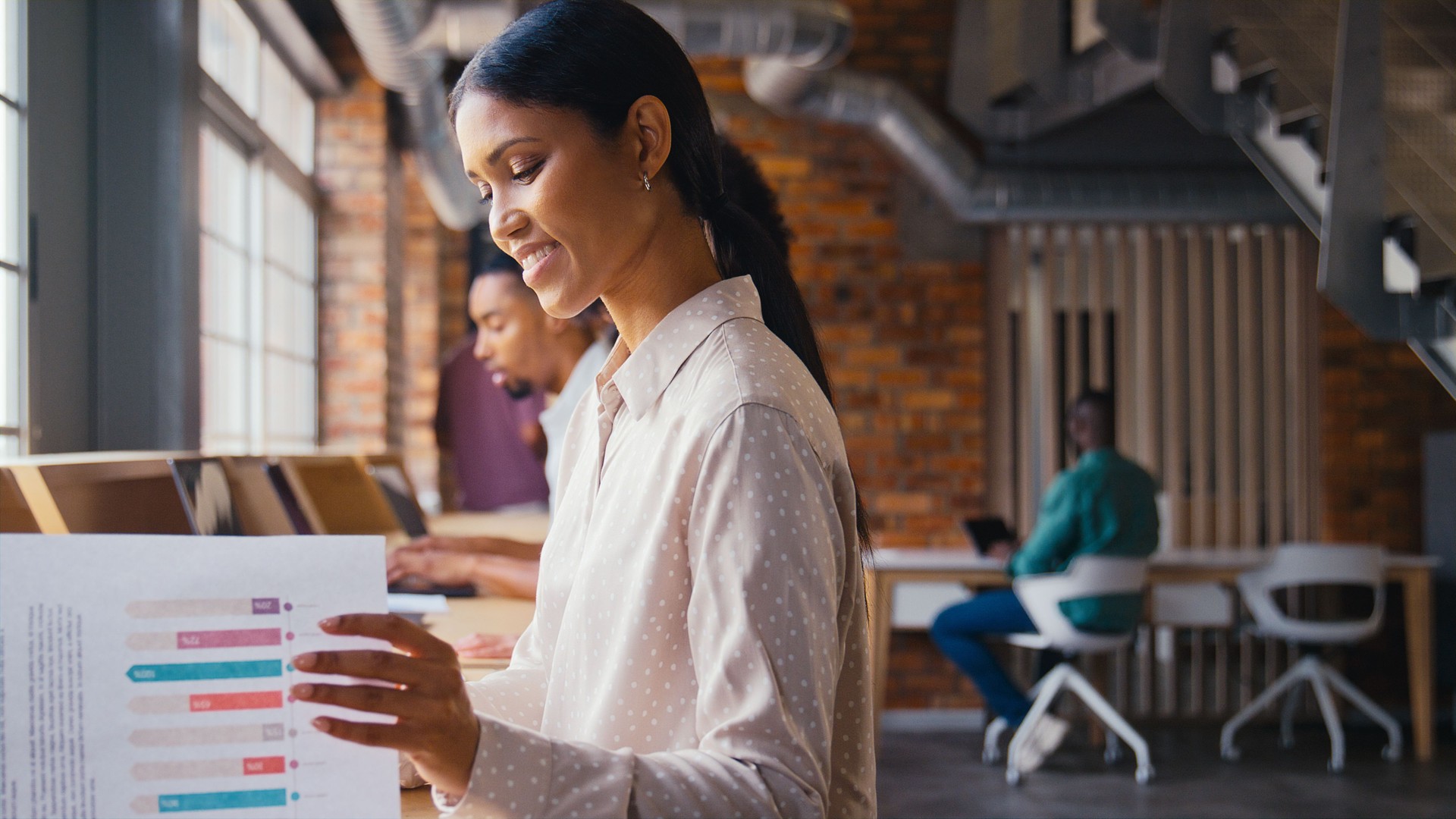 Businesswoman With Laptop Working In Busy Modern Multi-Cultural Open Plan Office Looking At Document