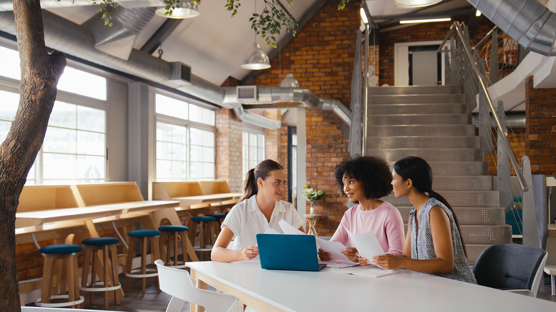 Three Young Businesswomen Meeting In Modern Office Sitting Around Table Working On Laptop Together